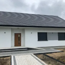 Nowoczesny dom jednorodzinny z białą elewacją, ciemnoszarą dachówką i brukowanym podjazdem w trakcie prac wykończeniowych ogrodu.