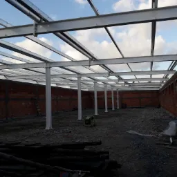 Wnętrze budowanej hali z widoczną stalową konstrukcją szkieletową, częściowo wymurowanymi ścianami z cegły dziurawki i generatorem prądu pośrodku.