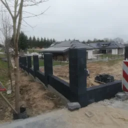 Wznoszony, nowoczesny, czarny ogrodzenie modułowe na posesji z domami jednorodzinnymi w tle, widoczna tablica reklamowa firmy ochroniarskiej.