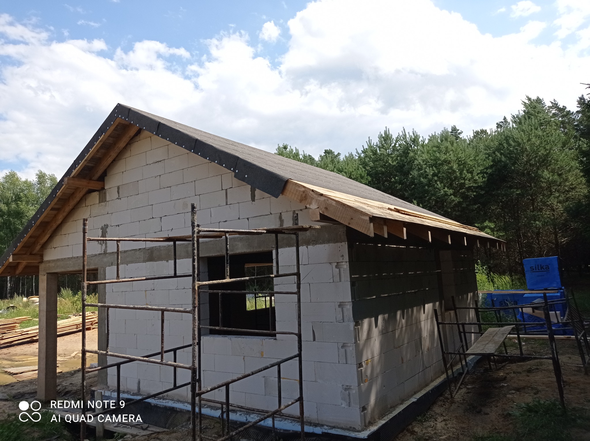 Niedokończony budynek z białych bloczków, częściowo pokryty czarnym materiałem dachowym, widoczne rusztowanie i drewniane elementy konstrukcji dachu, w tle las iglasty i błękitne niebo z chmurami.