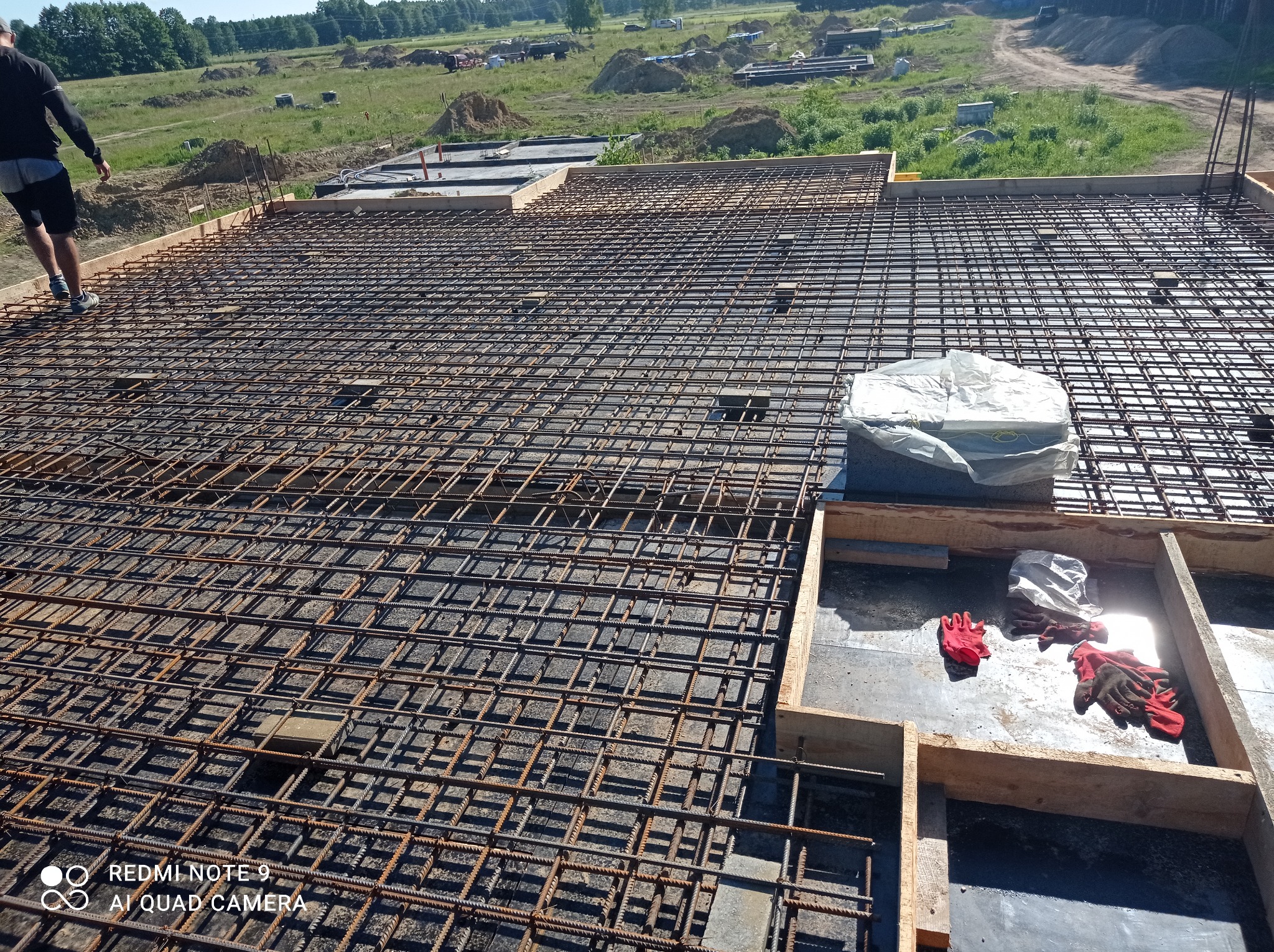 Zbrojenie płyty fundamentowej z widocznymi prętami zbrojeniowymi, szalunkiem drewnianym, rękawicami roboczymi i osobą w oddali na tle krajobrazu budowlanego.