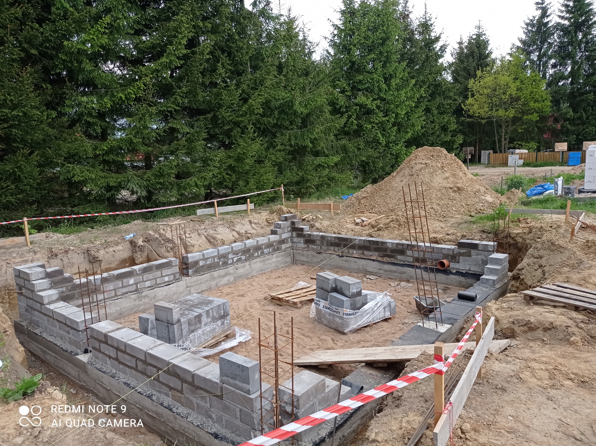 Fundamenty domu w trakcie budowy, widoczne bloczki betonowe, zbrojenie, wykop ziemny, teren budowy zabezpieczony taśmą ostrzegawczą, w tle drzewa.