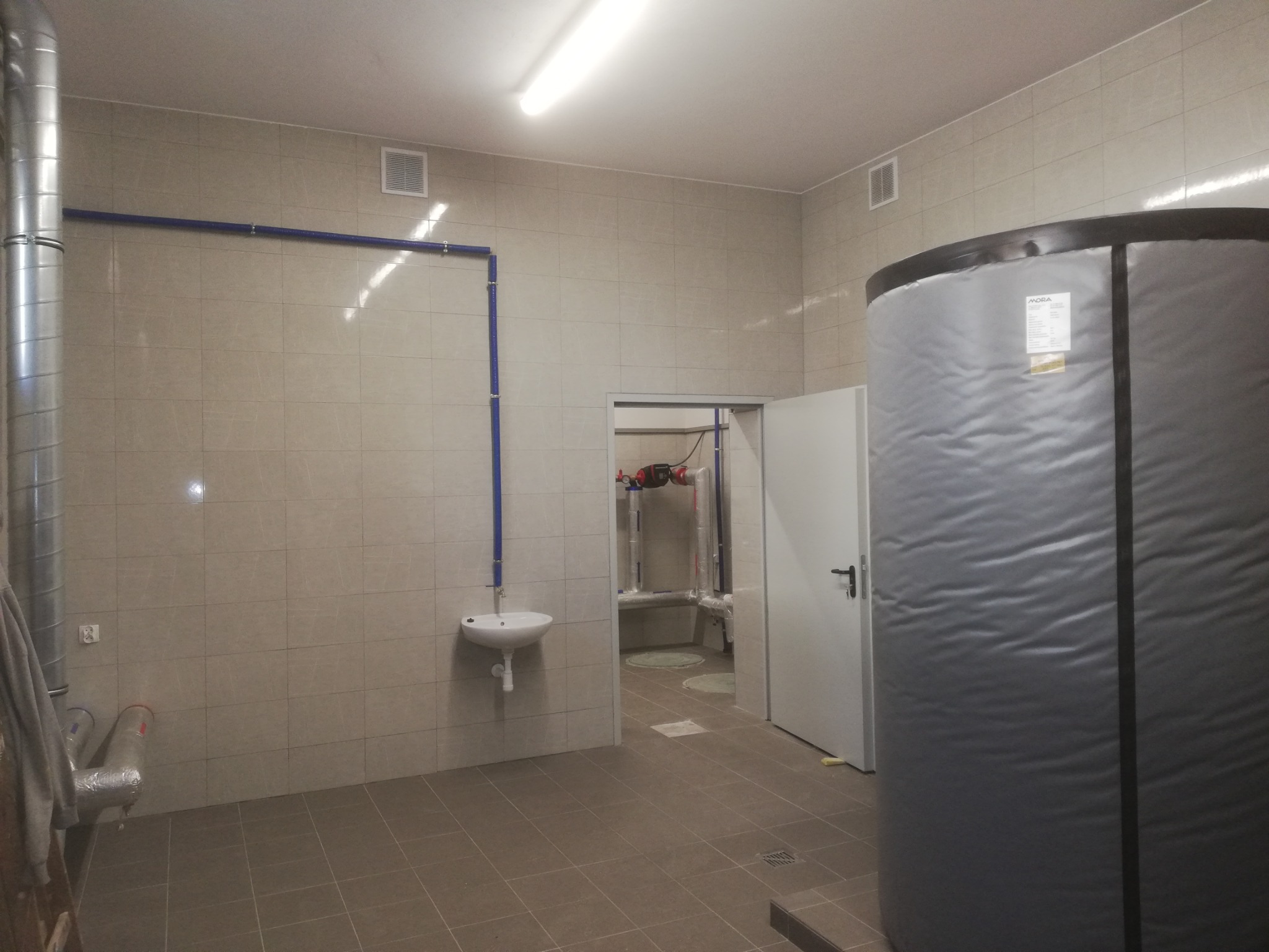 Wnętrze kotłowni z dużym zbiornikiem wody, instalacją hydrauliczną, umywalką i wyłożone płytkami ceramicznymi w odcieniach beżu.