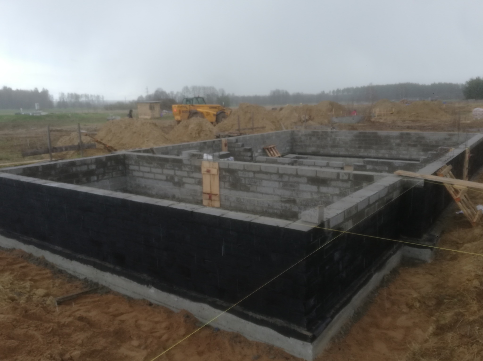 Budowa fundamentów domu z bloczków betonowych, zabezpieczonych czarną izolacją, na tle placu budowy z żółtą ładowarką teleskopową i stertami ziemi w pochmurny dzień.