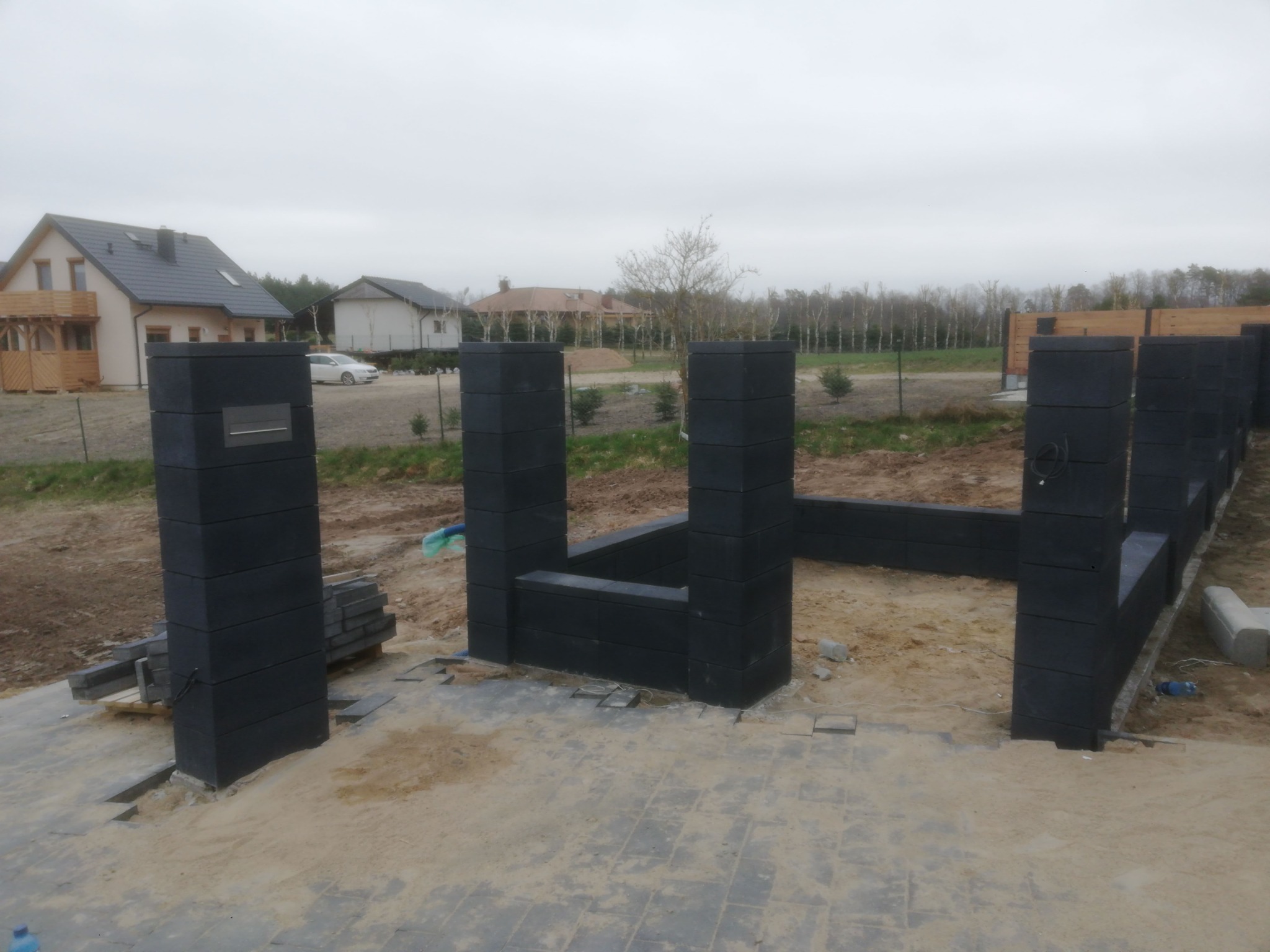 Budowa ogrodzenia: Czarny, modułowy system ogrodzeniowy w trakcie montażu, widoczne słupki i podmurówka, przygotowany teren z kostką brukową, domy w tle.