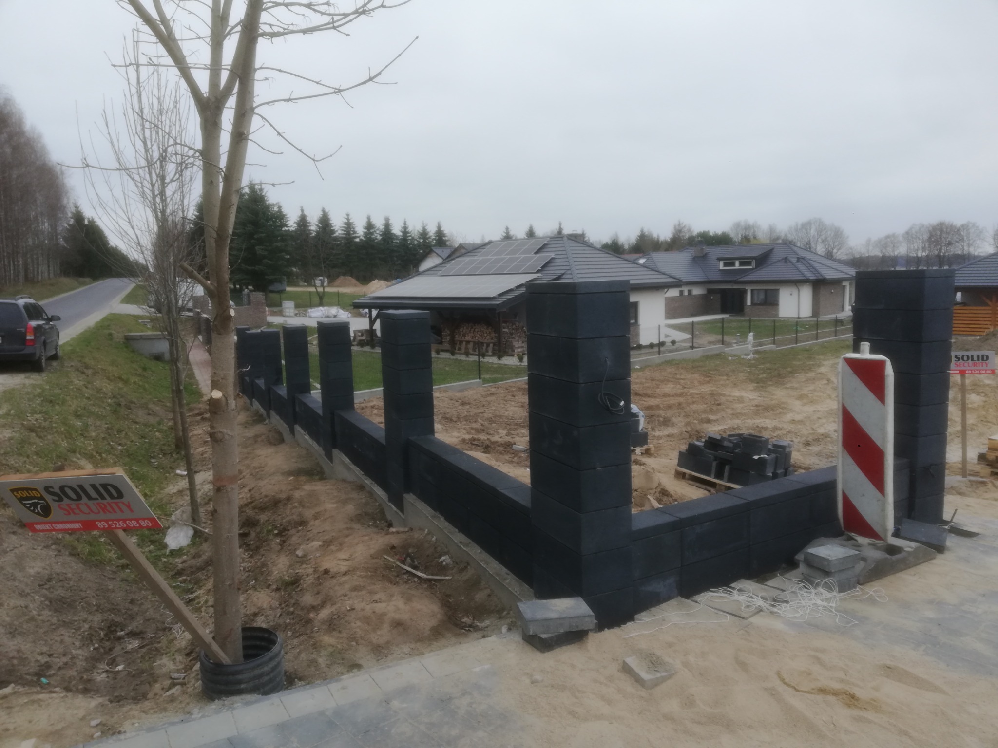 Wznoszony, nowoczesny, czarny ogrodzenie modułowe na posesji z domami jednorodzinnymi w tle, widoczna tablica reklamowa firmy ochroniarskiej.