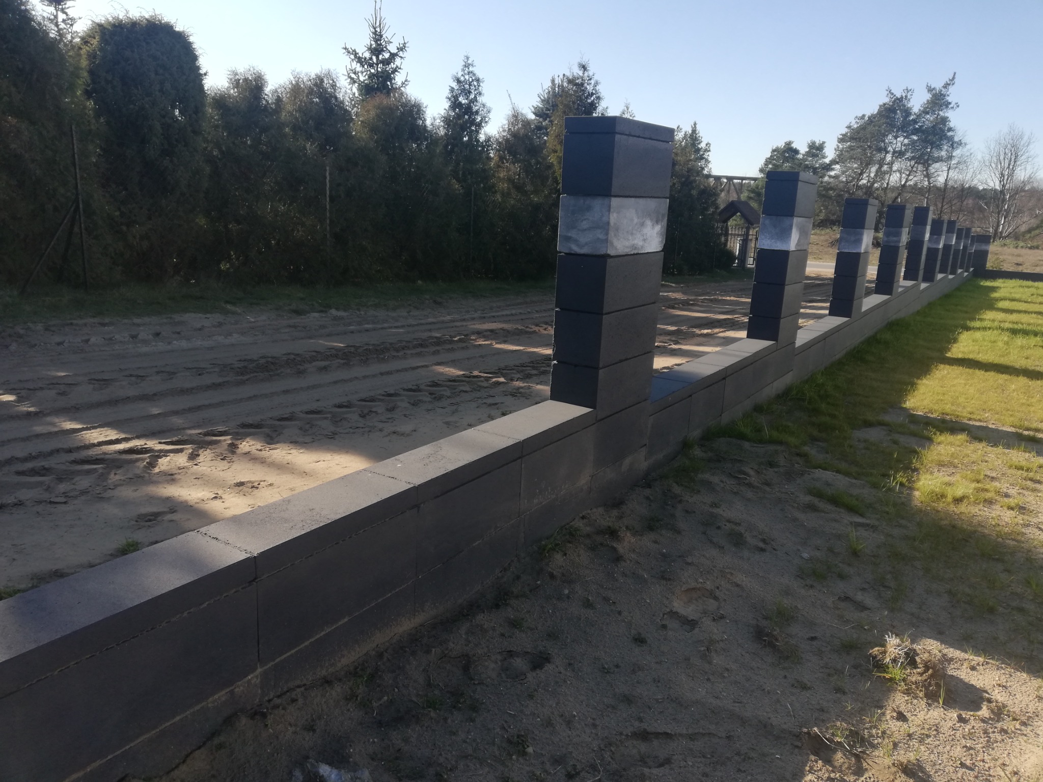 Nowoczesne ogrodzenie z szarych betonowych bloczków z elementami dekoracyjnymi w kolorze jasnego kamienia, widoczne na tle trawnika i drzew.