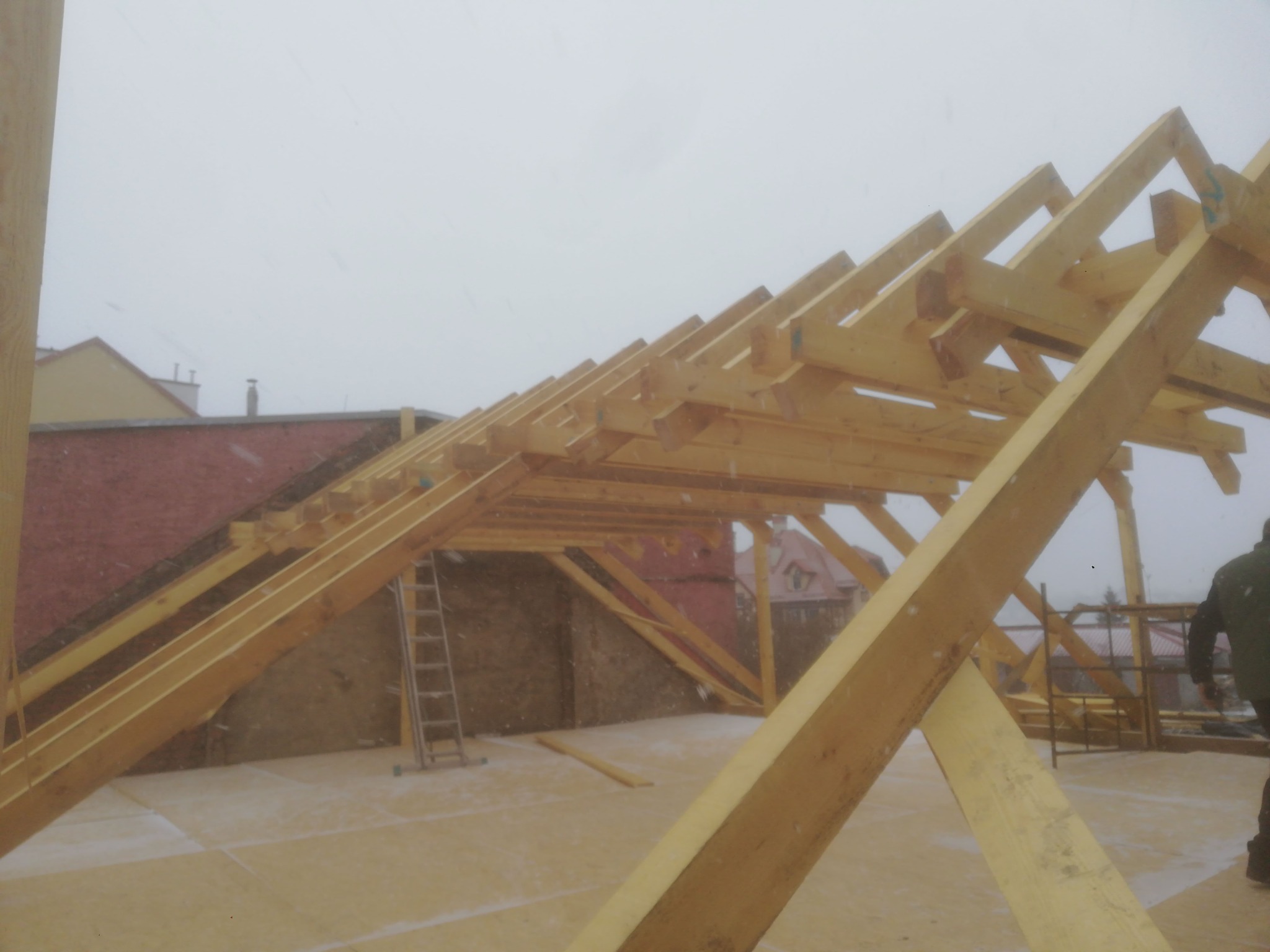 Konstrukcja drewniana dachu w trakcie budowy, widoczne krokwie i elementy więźby dachowej, w tle drabina i fragment budynku w zimowej scenerii.
