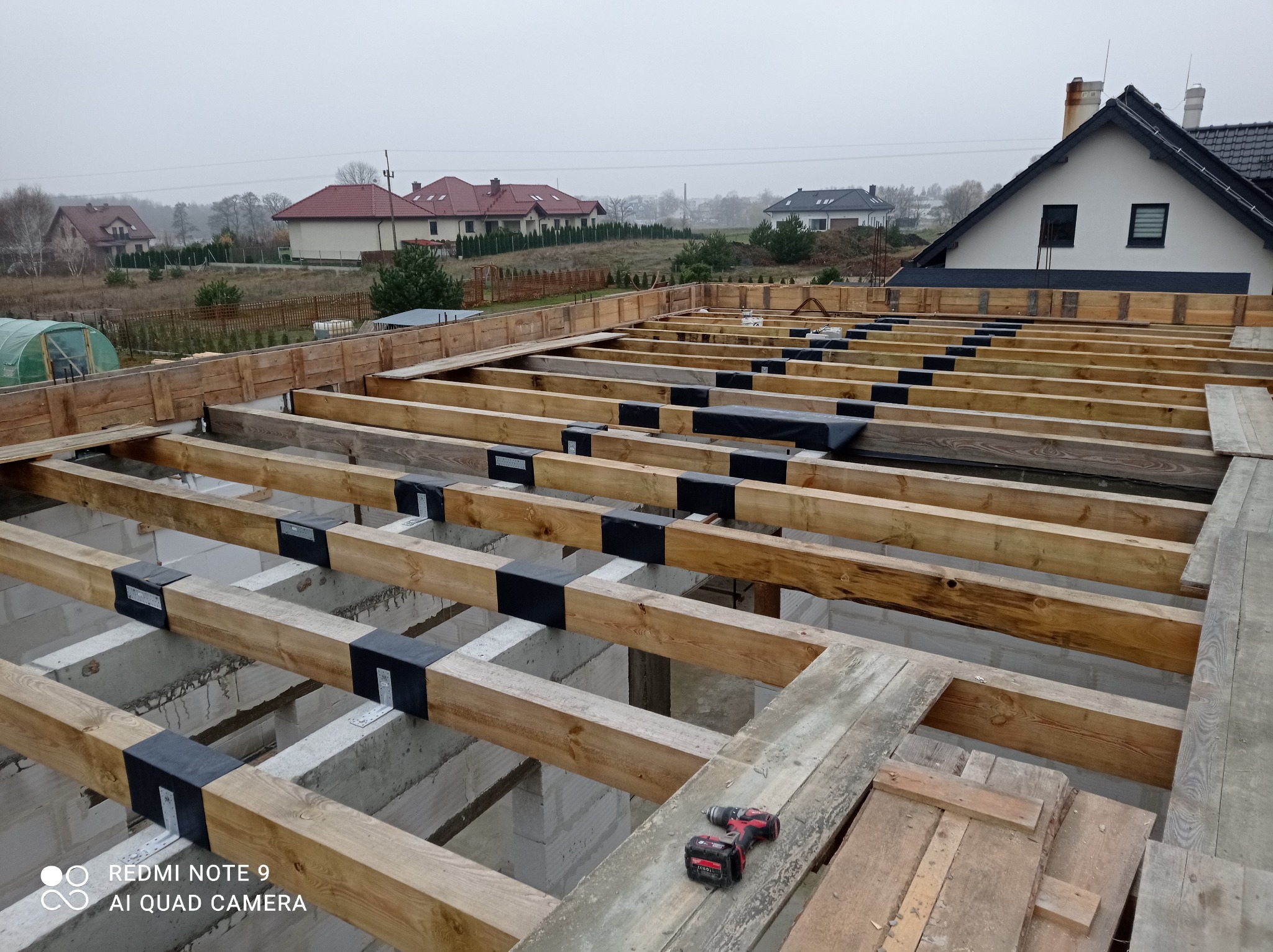 Konstrukcja drewniana stropu nad piwnicą w trakcie budowy, widoczne drewniane belki połączone metalowymi łącznikami, w tle domy jednorodzinne w mglisty dzień, w kadrze wiertarko-wkrętarka...