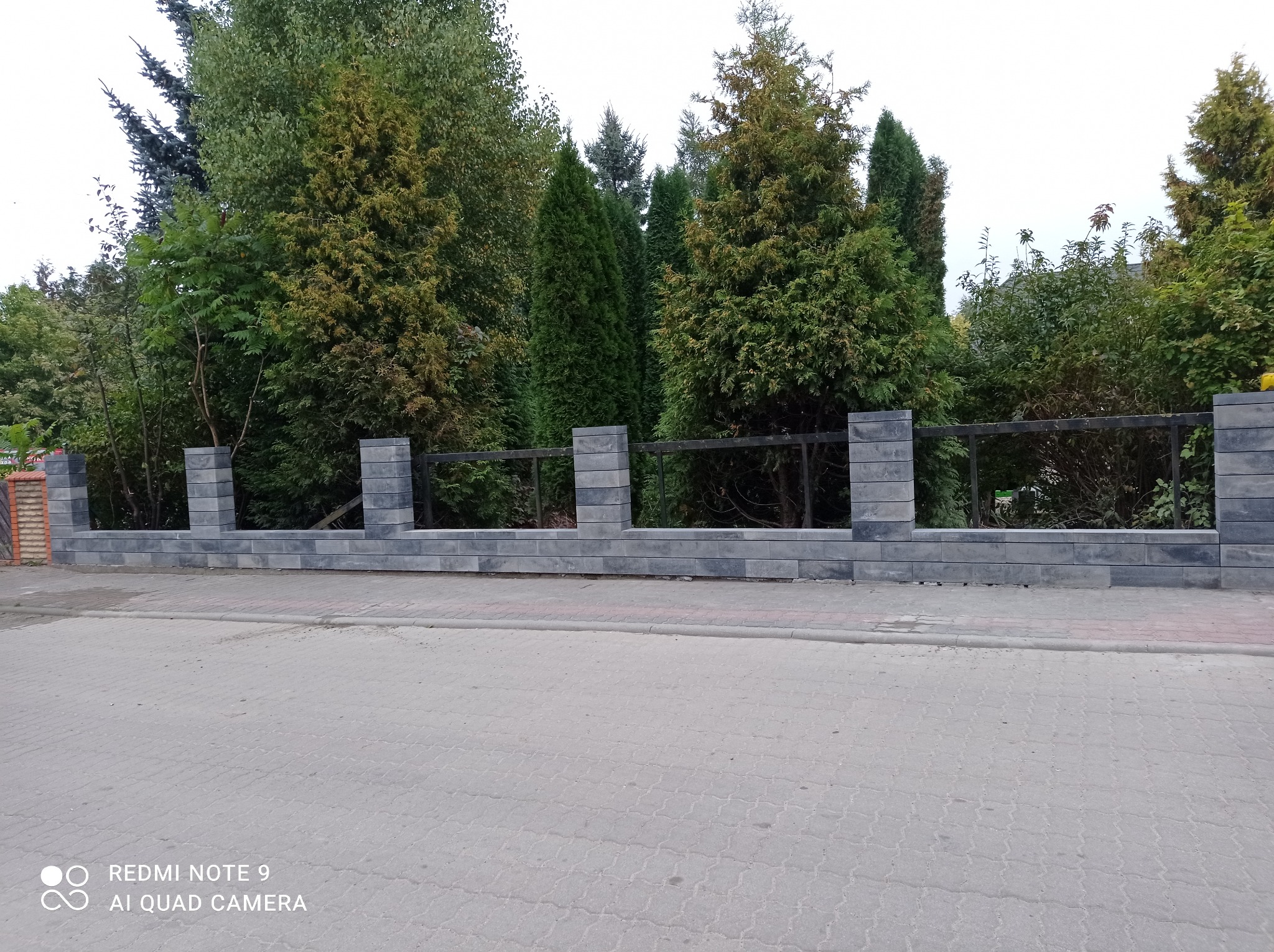 Nowoczesne ogrodzenie z szarej kostki betonowej z metalowymi przęsłami, widok z ulicy na tle zieleni.