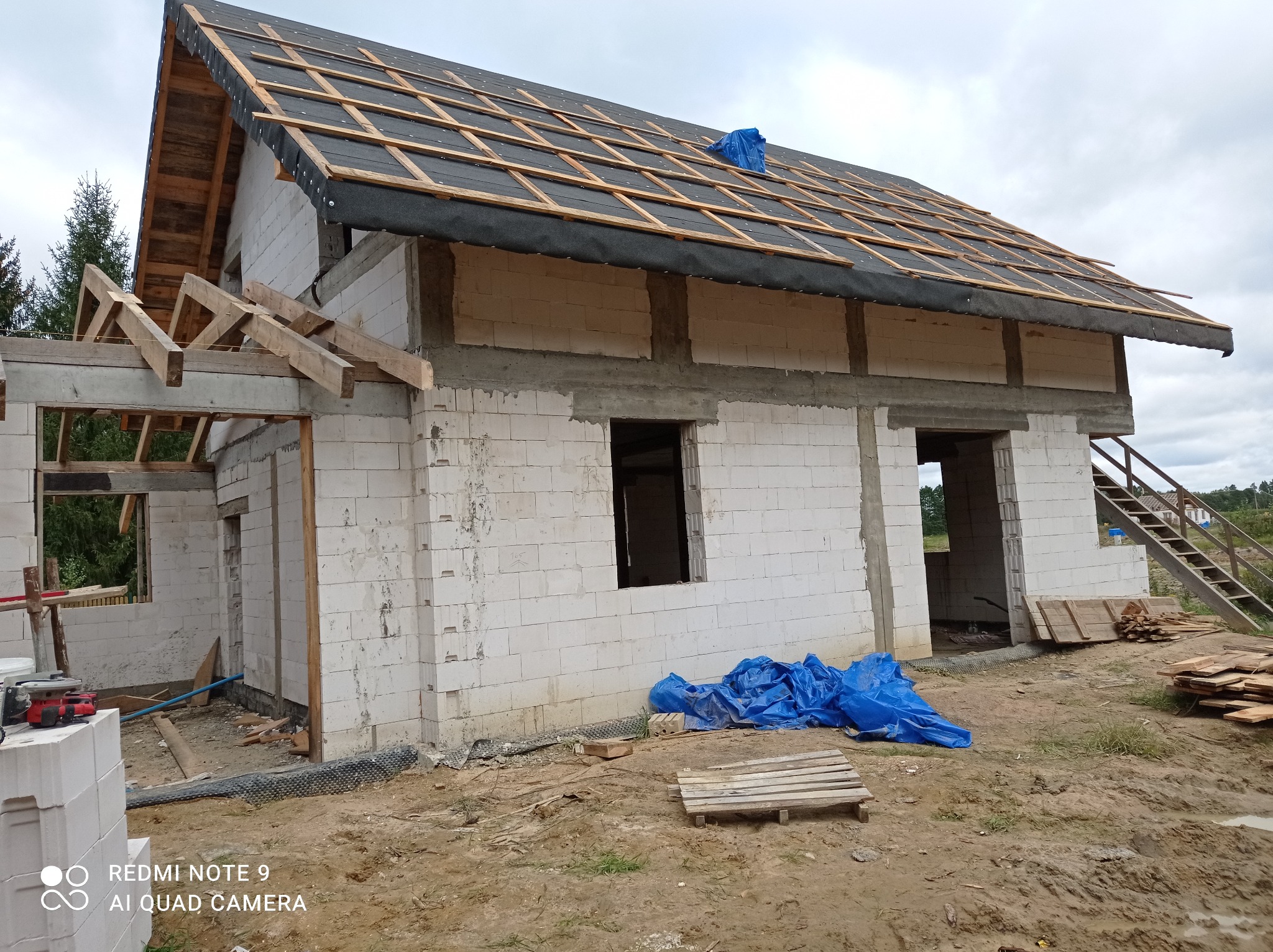 Surowy stan domu w trakcie budowy z widocznym dachem pokrytym membraną i łatami, ściany z białego bloczka, niezadaszona część z drewnianą konstrukcją, niebieska plandeka na dachu i ziemi, oraz stos...