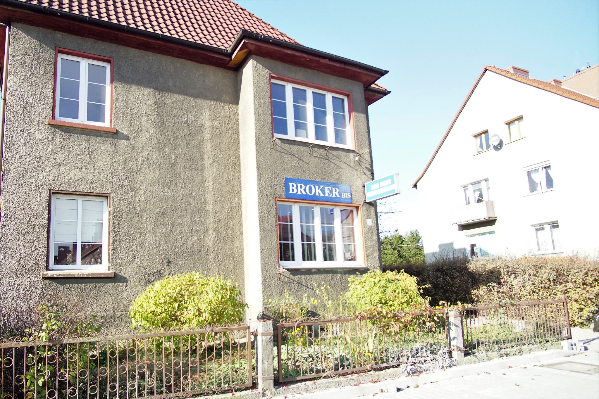 Budynek biura nieruchomości Broker Bis z szyldem, widoczny z zewnątrz, z ozdobnym ogrodzeniem i krzewami przed wejściem.