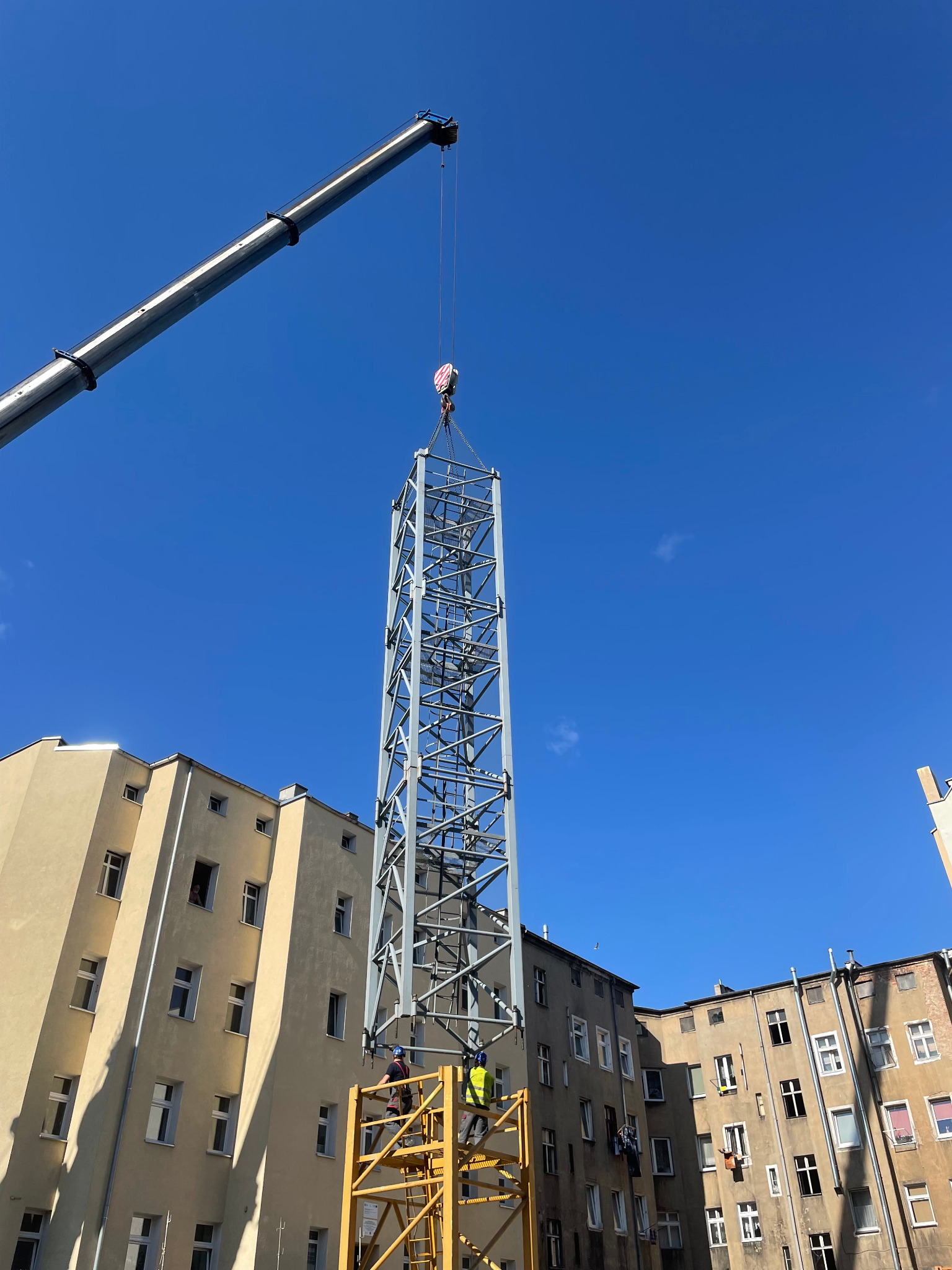 Montaż metalowej konstrukcji szkieletowej za pomocą dźwigu na tle budynków mieszkalnych i błękitnego nieba, pracownicy na żółtym rusztowaniu w trakcie pracy.