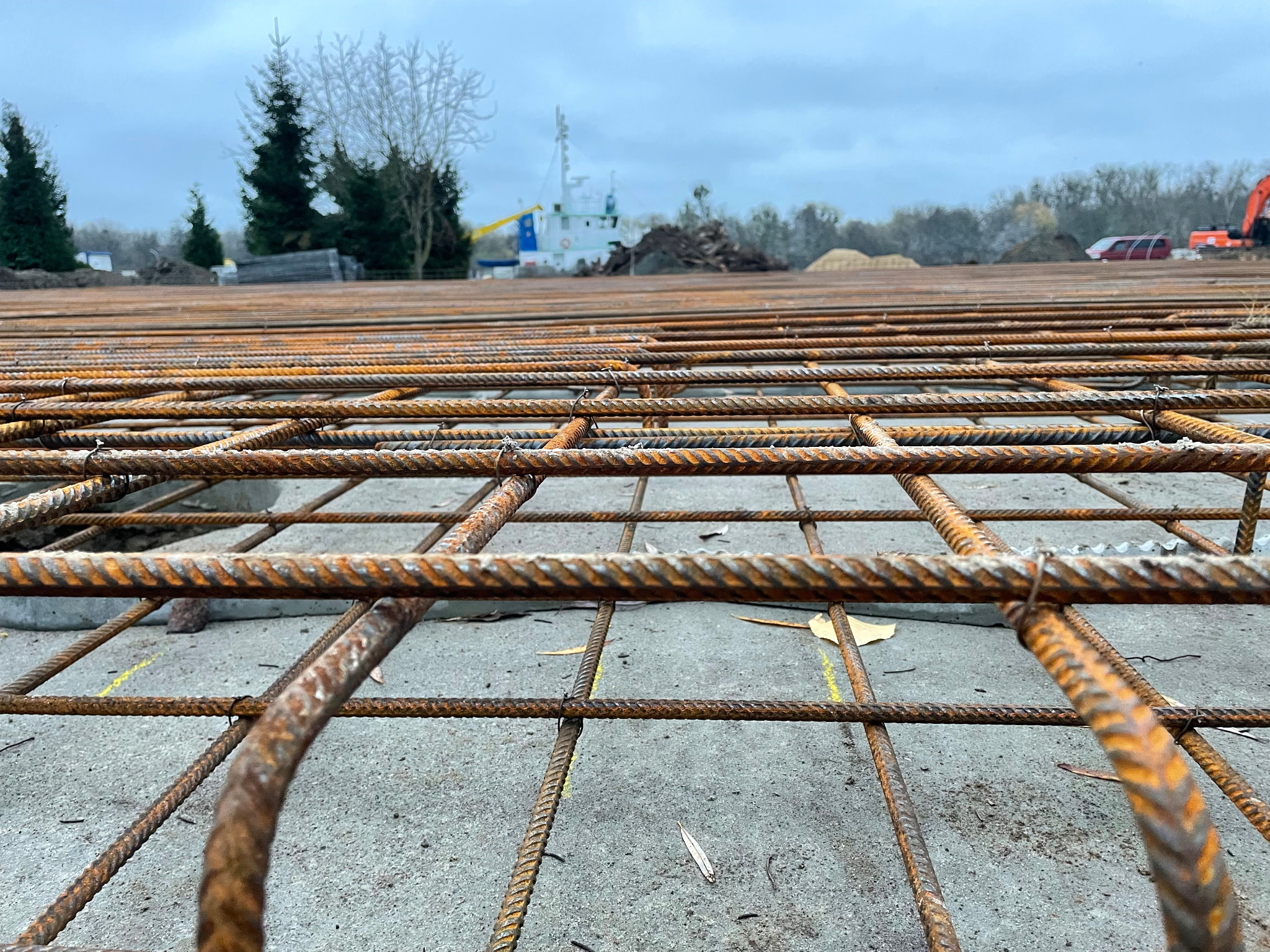 Siatka zbrojeniowa z widoczną rdzą na prętach, ułożona na betonowej powierzchni z żółtymi oznaczeniami, w tle zieleń, statek i sprzęt budowlany pod pochmurnym niebem.