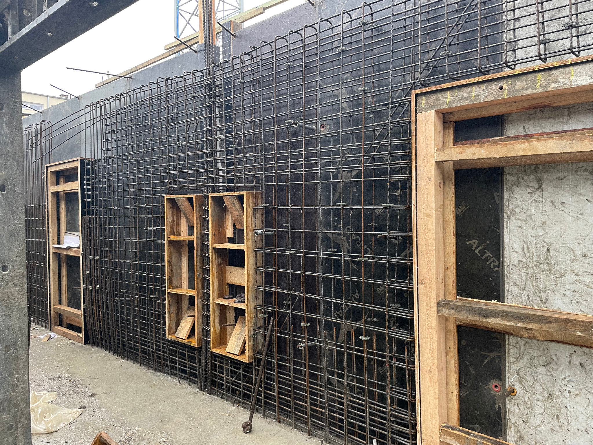 Zbrojenie pionowej ściany szalunkowej z drewnianymi otworami okiennymi, widoczne elementy mocujące i fragment żurawia w tle.