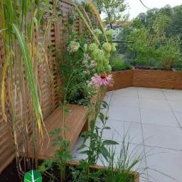 Ogród na balkonie z drewnianą ławką wbudowaną w donicę, ozdobne trawy i kwitnące rośliny na tle drewnianej kratownicy i zieleni drzew.