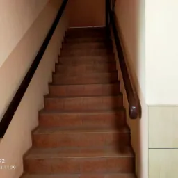 Klatka schodowa z brązowymi płytkami w budynku w Lubinie, widok z dołu na górę schodów, z ciemnymi poręczami po bokach, ściany w odcieniach beżu i kremu.