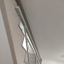 Wnętrze klatki schodowej z widoczną ścianą wykończoną tynkiem mozaikowym w odcieniach beżu i brązu, białą balustradą metalową i fragmentem białego sufitu