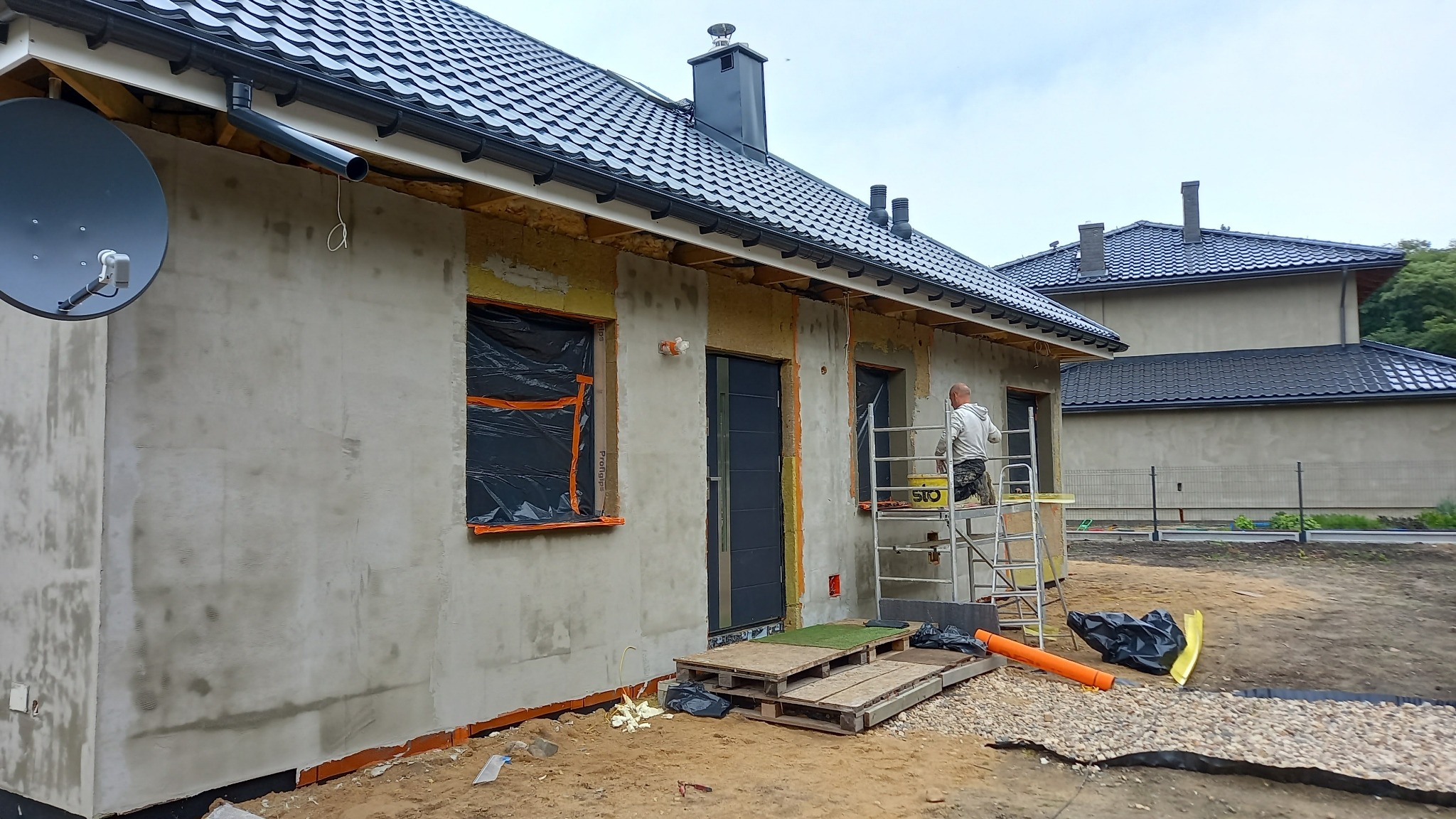 Dom jednorodzinny w trakcie nakładania warstwy elewacyjnej: widoczne okna zabezpieczone folią, fragment ocieplenia, rusztowanie z pracownikiem i przygotowane podłoże.