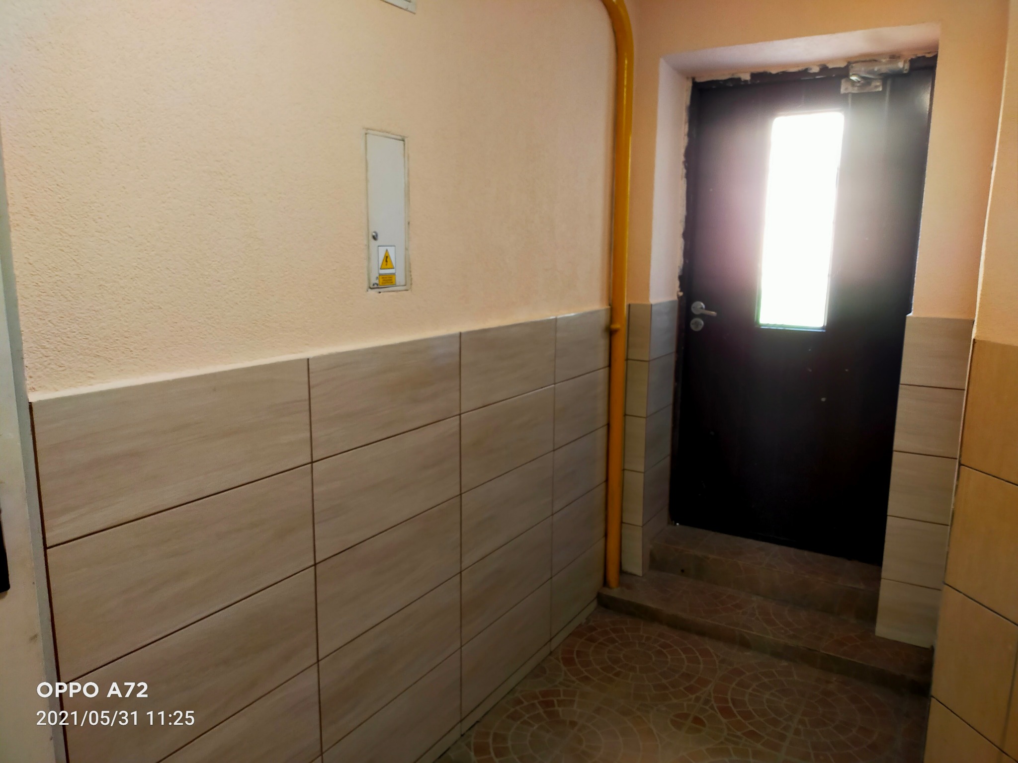 Wnętrze klatki schodowej z wykończoną ścianą w płytkach imitujących drewno, widoczna rura gazowa i ciemne drzwi z przeszkleniem, podłoga wyłożona okrągłymi płytkami.