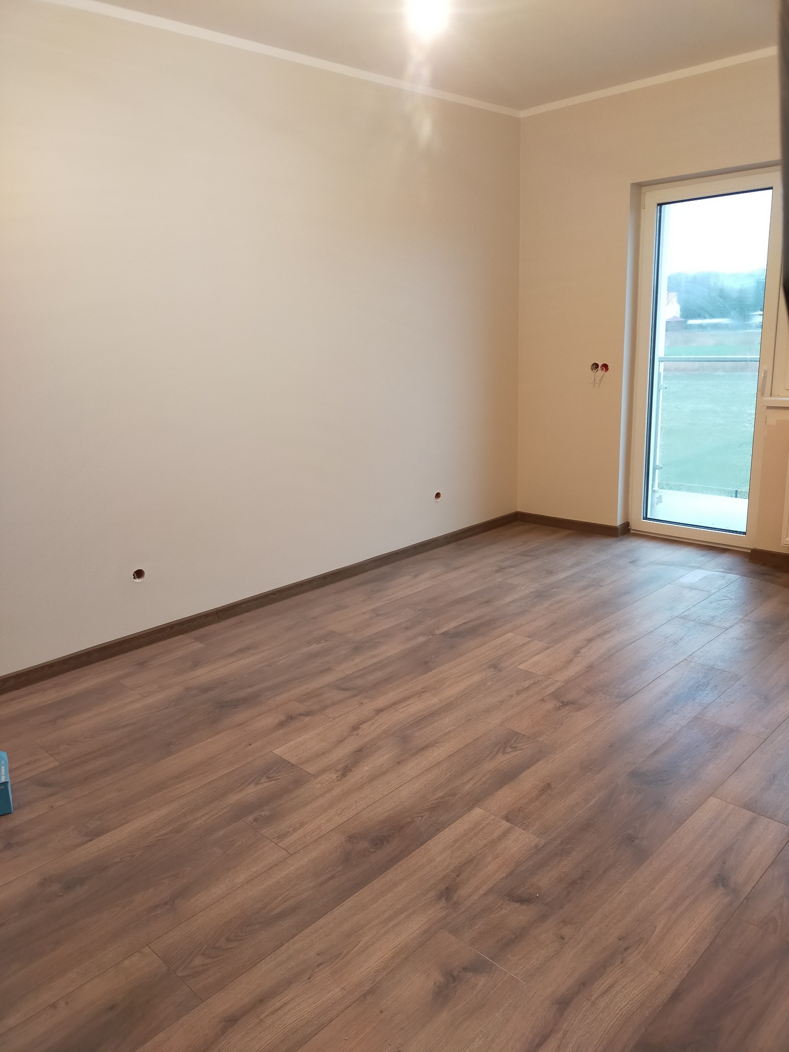 Pusty pokój z panelami podłogowymi w odcieniach brązu, świeżo pomalowane ściany w kolorze ecru z widocznymi otworami na gniazdka elektryczne i parapet okienny z widokiem na zieleń za oknem.