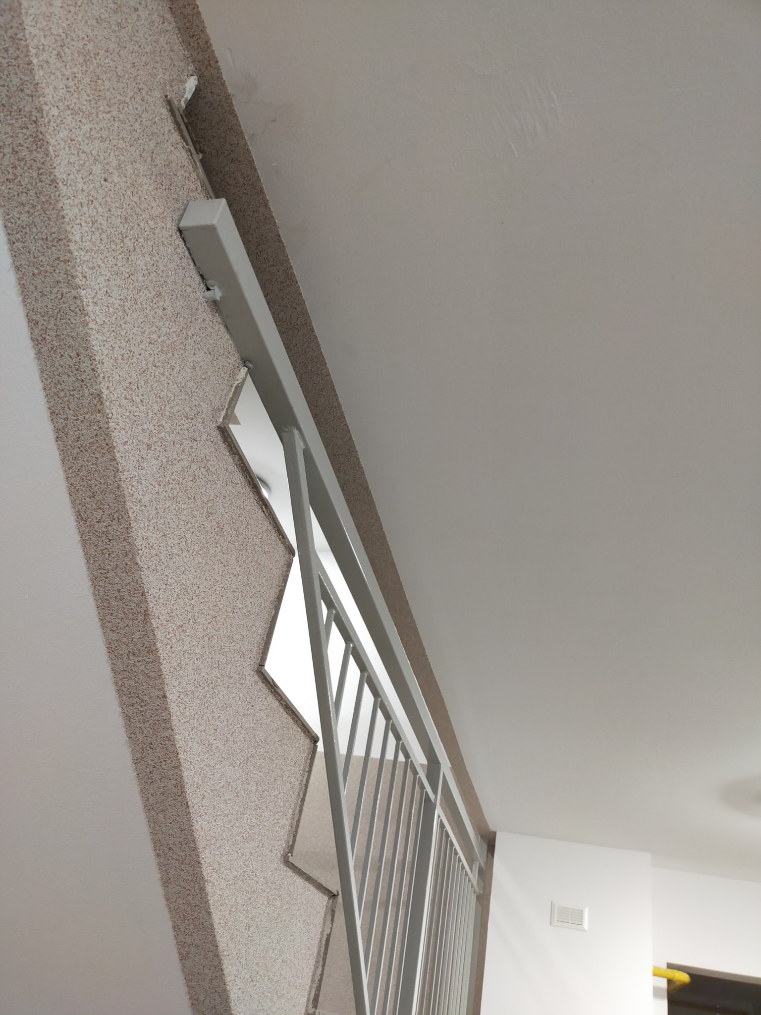 Wnętrze klatki schodowej z widoczną ścianą wykończoną tynkiem mozaikowym w odcieniach beżu i brązu, białą balustradą metalową i fragmentem białego sufitu