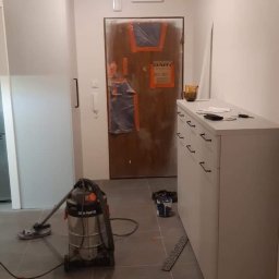 MMyrcha - Korytarz w trakcie remontu: zabezpieczone drzwi wejściowe, odkurzacz przemysłowy, pomalowane ściany i szare płytki podłogowe.