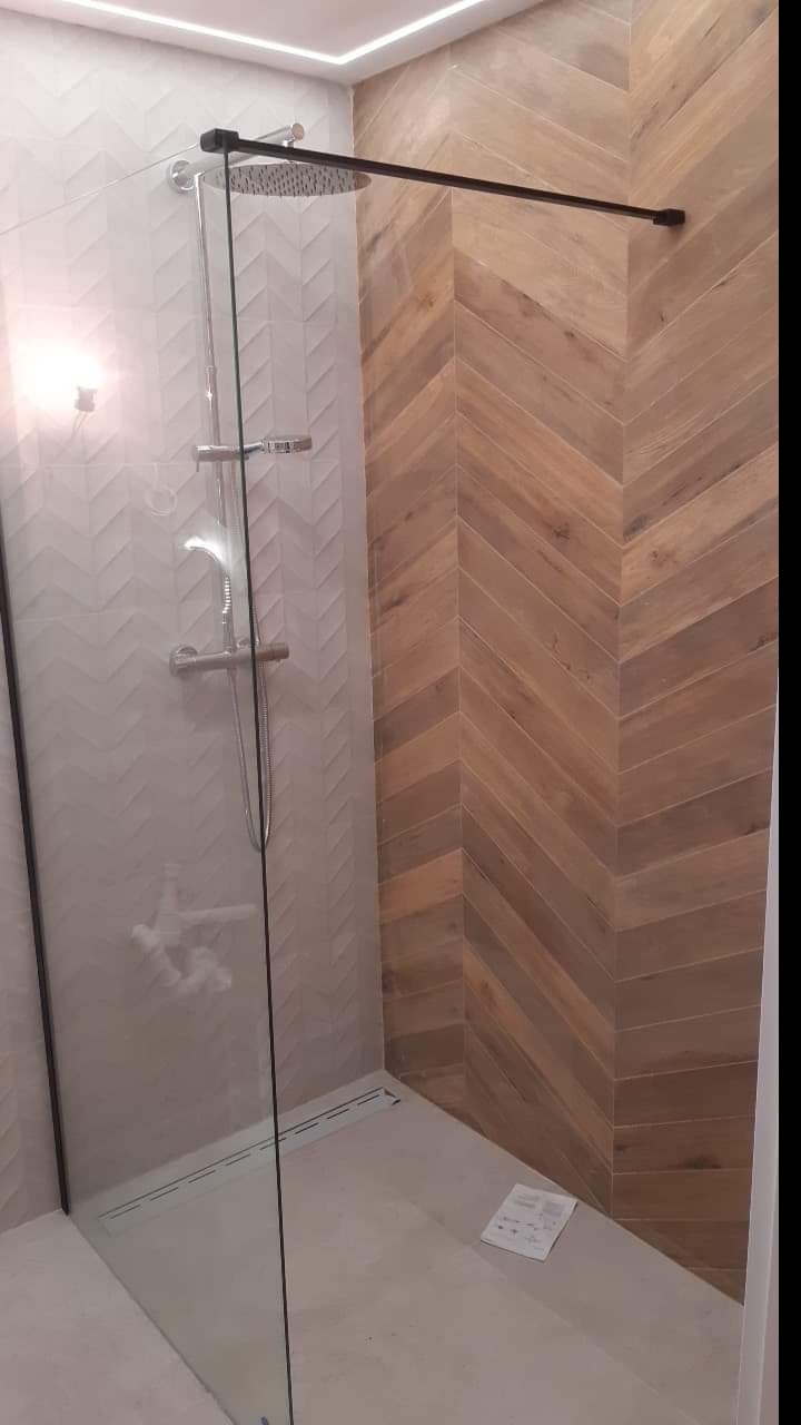 Nowoczesna kabina prysznicowa z deszczownicą i szklaną ścianką, wykończona płytkami w dwóch odcieniach: białym z subtelnym wzorem jodełki i brązowym imitującym drewno, ułożonym w jodełkę. Dodatkowo...