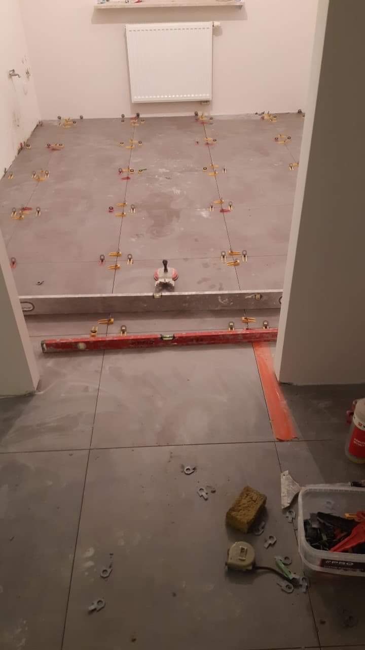 Układanie szarych płytek podłogowych w trakcie prac, widoczne poziomice, krzyżyki dystansowe, gąbka, miarka zwijana i pojemnik z narzędziami.