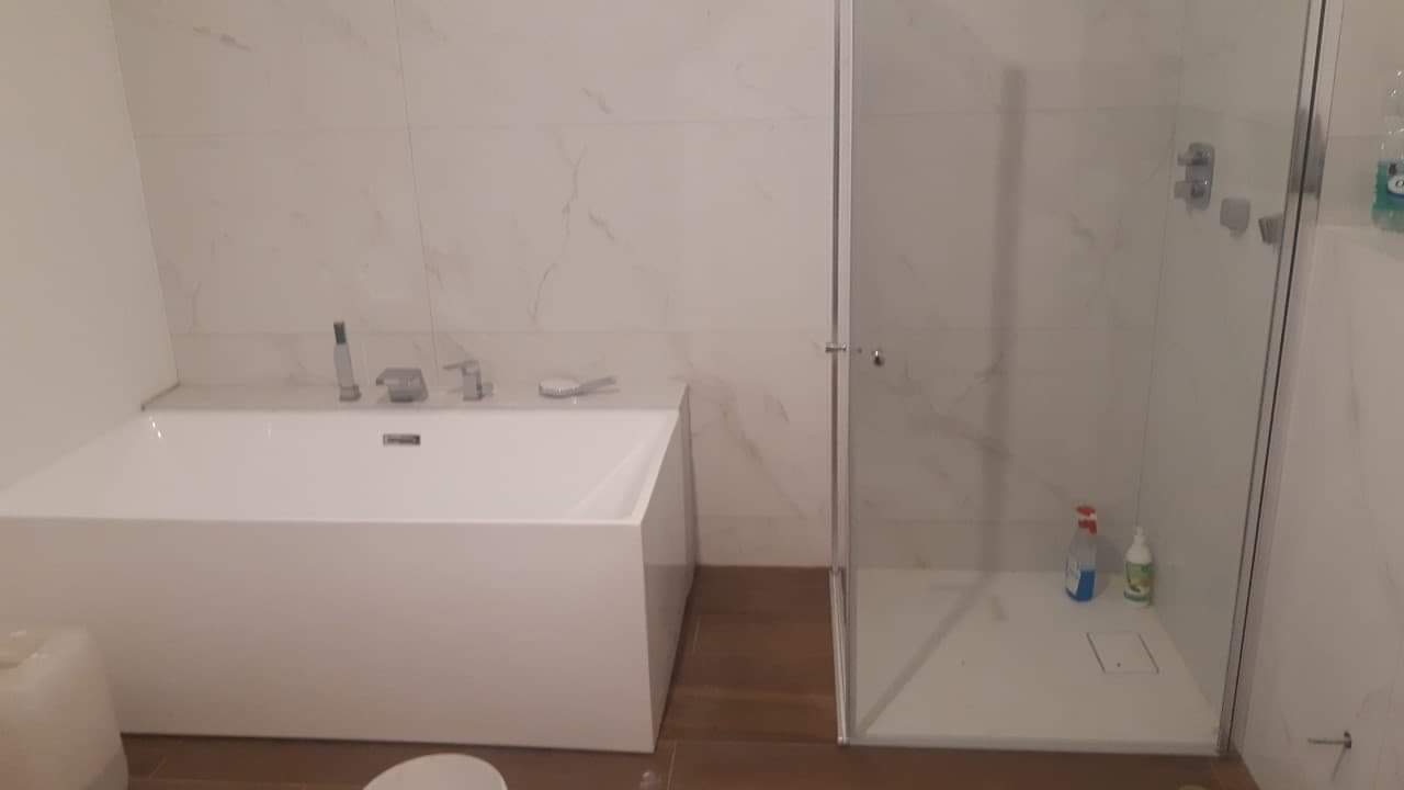 Nowoczesna łazienka z białą, prostokątną wanną i kabiną prysznicową typu walk-in, wykończona płytkami imitującymi marmur, z drewnopodobną podłogą i widocznymi środkami czystości w kabinie.