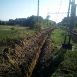 Długi, prosty wykop w ziemi biegnący wzdłuż torów kolejowych, otoczony zielenią i polami.