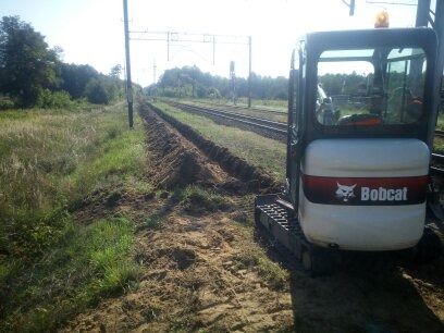 Mini koparka Bobcat na torach kolejowych po wykopaniu wąskiego rowu wzdłuż torów, widoczne słupy i trawa. Rów wykopany na potrzeby instalacji.