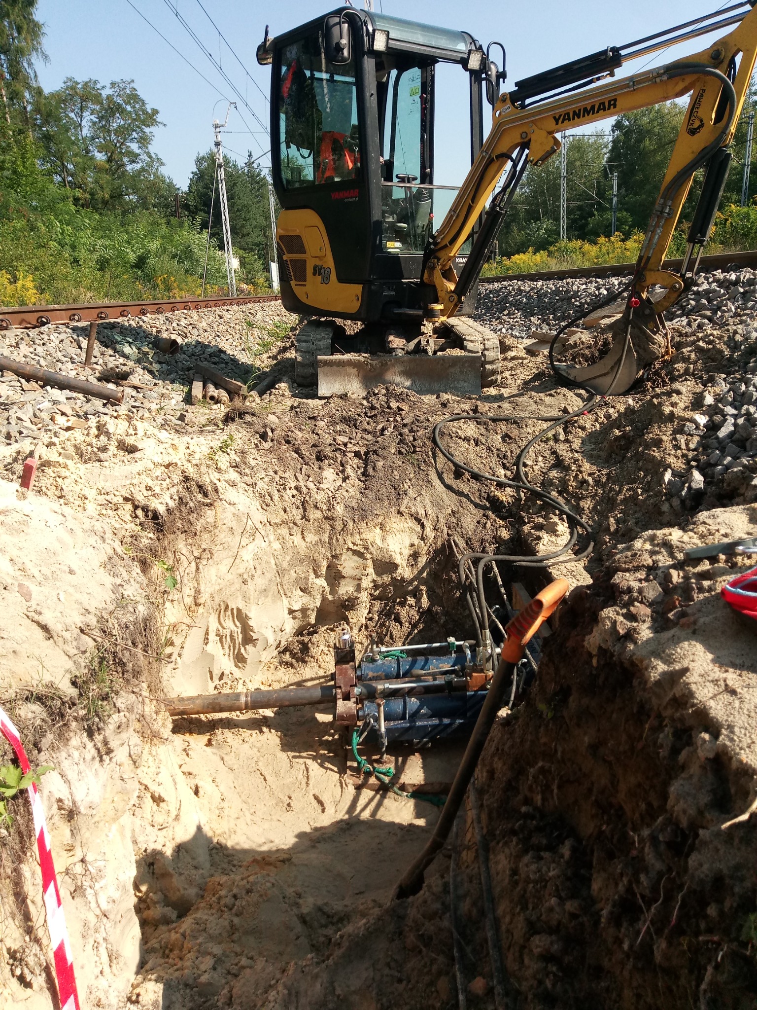 Żółta minikoparka Yanmar stoi obok wykopu z widocznym urządzeniem do przewiertów pod torami kolejowymi, zabezpieczonymi taśmą ostrzegawczą.