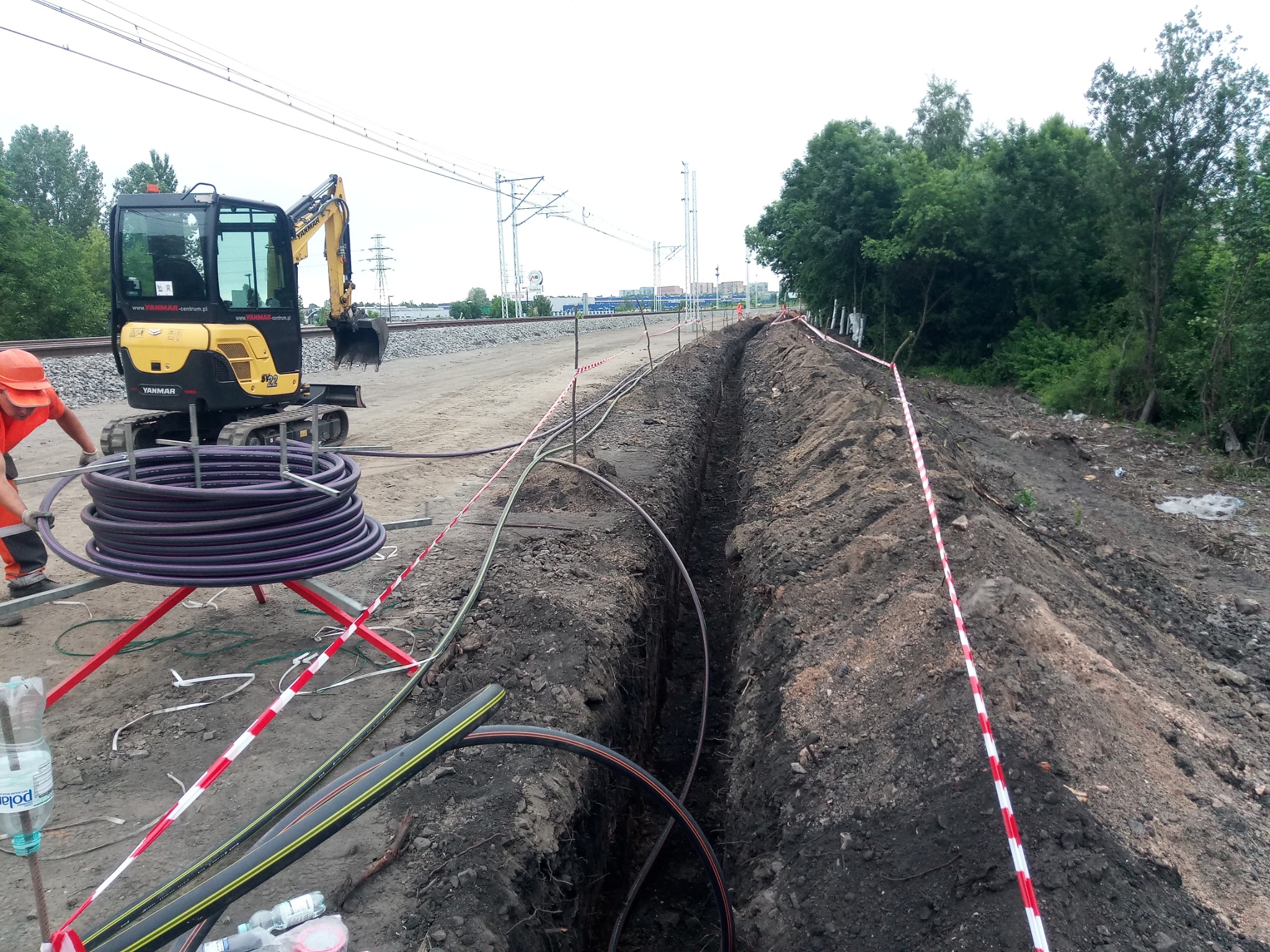 Żółta mini koparka Yanmar obok wykopanego wąskiego rowu, zabezpieczonego taśmą ostrzegawczą, z widocznymi kablami wewnątrz i zwojem rur na stojaku, w tle tor kolejowy i zieleń.