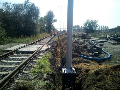 Wykop wąskoprzestrzenny wzdłuż torów kolejowych, widoczni pracownicy w odblaskowych kamizelkach, niebieski wąż odprowadzający wodę, teren budowy z gruzem w tle.