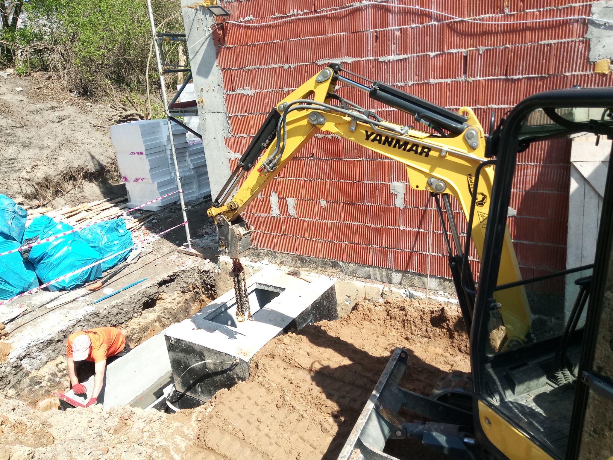 Żółty minikoparka Yanmar opuszcza betonowy element do wykopu, pracownik w pomarańczowej koszulce i czerwonych rękawicach wyrównuje podłoże, w tle mur z czerwonej cegły i niebieskie worki...