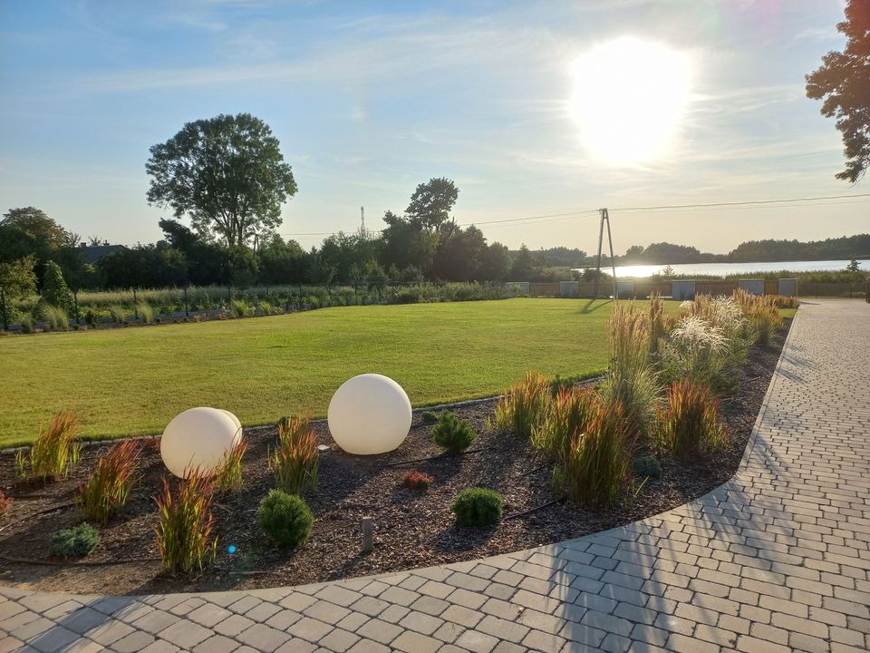 Starannie utrzymany trawnik z dekoracyjnymi trawami i kulistymi lampami ogrodowymi, widok na jezioro w tle, oświetlony zachodzącym słońcem, ścieżka z kostki brukowej.