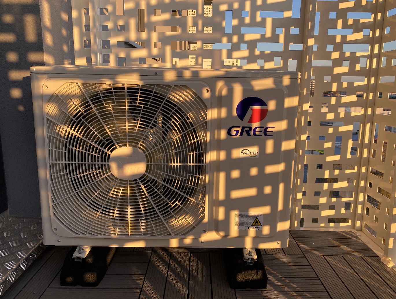 Zewnętrzna jednostka klimatyzacji marki Gree, zamontowana na balkonie, oświetlona popołudniowym słońcem, z widoczną ażurową osłoną. Na obudowie naklejka ostrzegawcza.