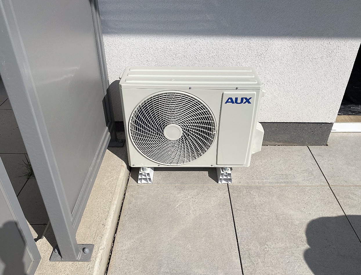 Zewnętrzna jednostka klimatyzacji AUX umieszczona na betonowym tarasie, obok szarej osłony; widoczne białe ściany budynku w tle.