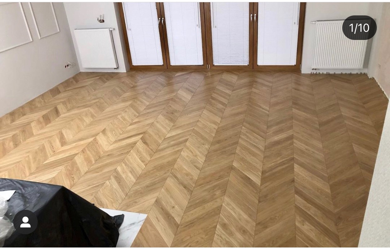 Jasny parkiet ułożony we wzór jodełki francuskiej w przestronnym pokoju z białymi ścianami i grzejnikami, widoczne drzwi balkonowe z żaluzjami.