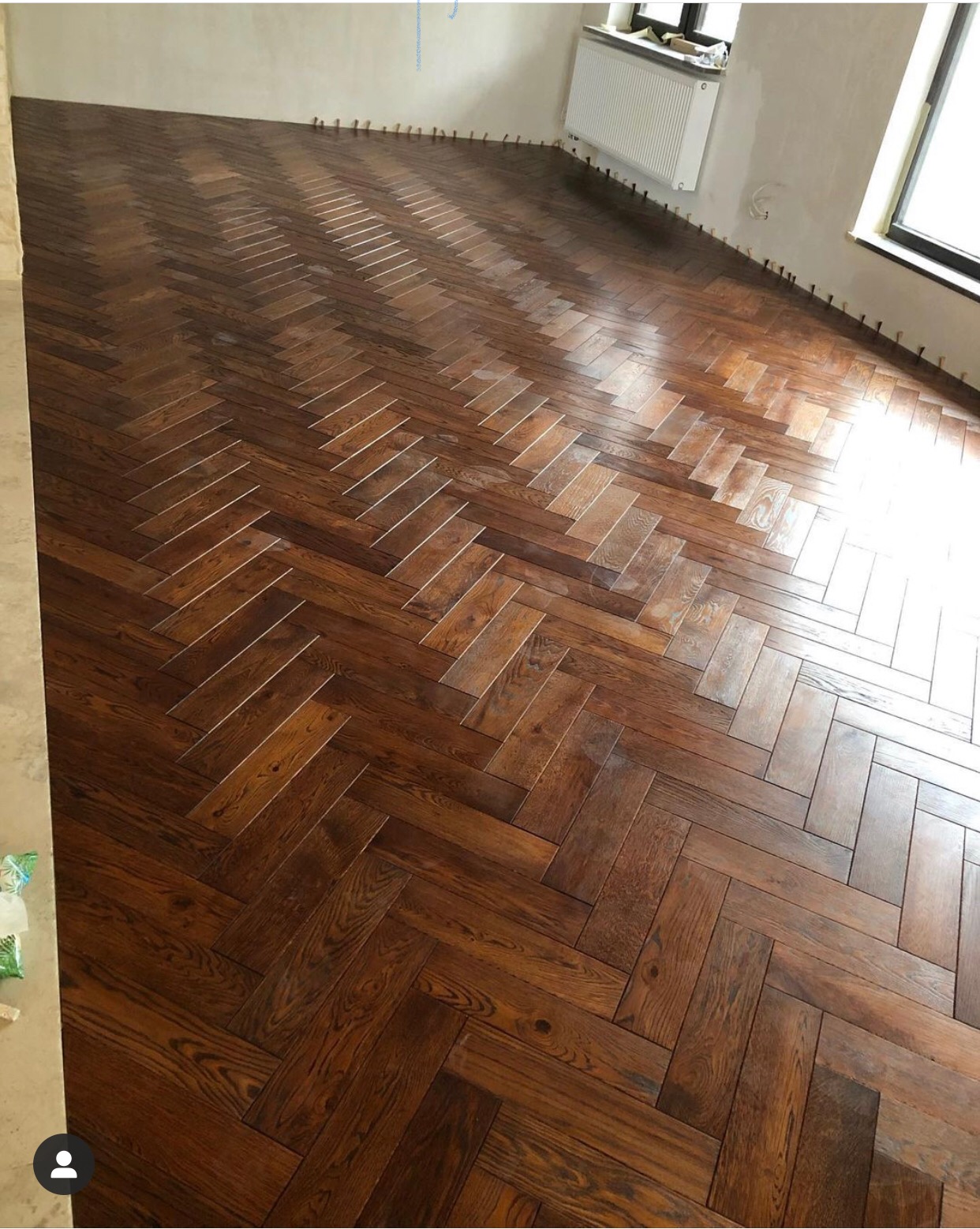 Ułożony parkiet w jodełkę z ciemnego drewna, widoczne kołki dystansowe przy ścianach, w tle okno i grzejnik.