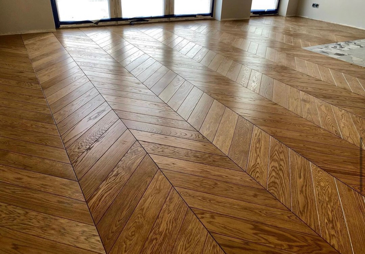 Ułożony parkiet w jodełkę francuską z naturalnego, lakierowanego drewna dębowego, widoczne okno z białymi ramami i fragment niedokończonej podłogi z szarymi płytkami.