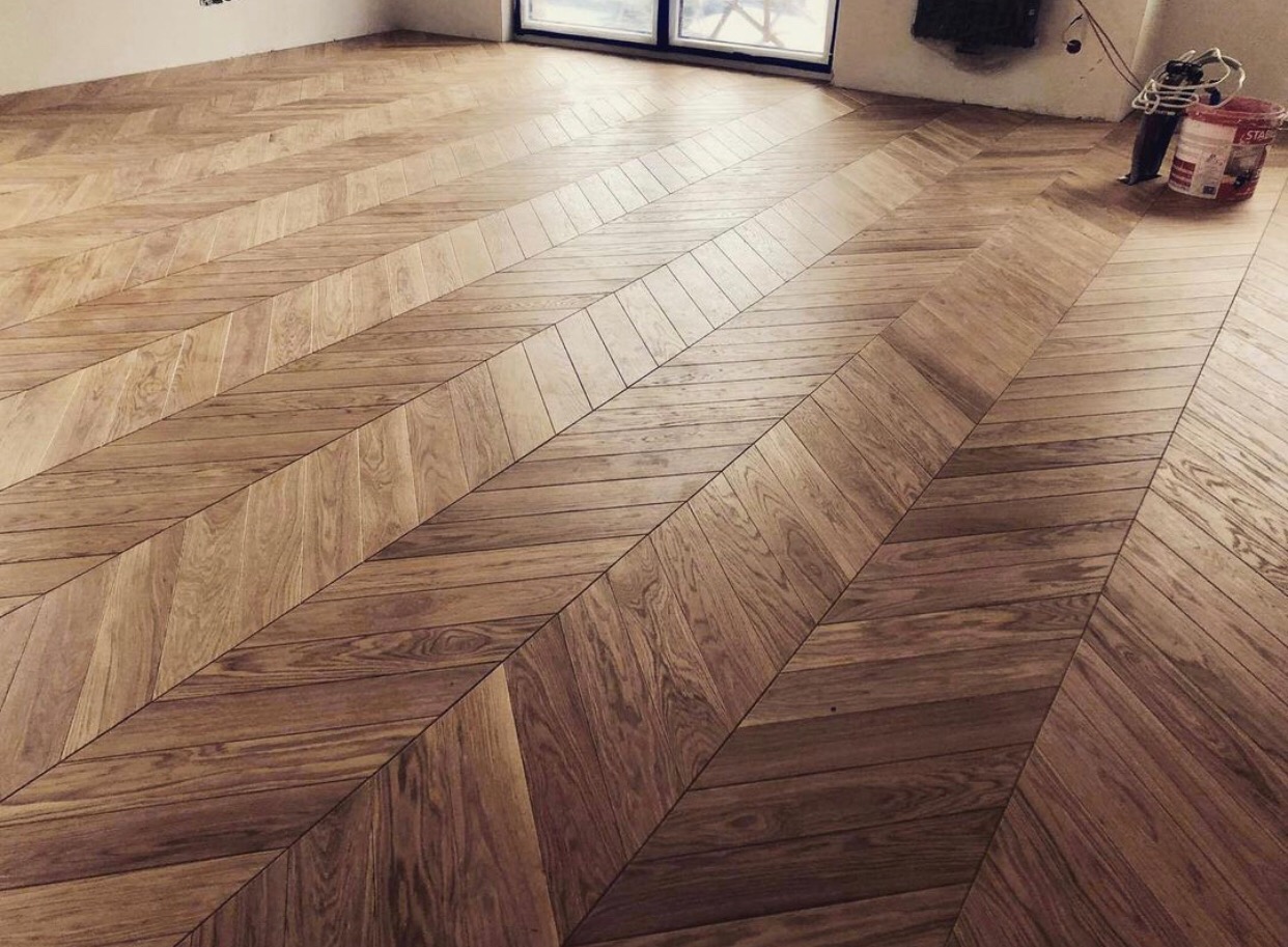 Ukończona podłoga z parkietu ułożonego we wzór jodełki francuskiej, widoczny fragment ściany i okno, w rogu wiaderko z narzędziami.