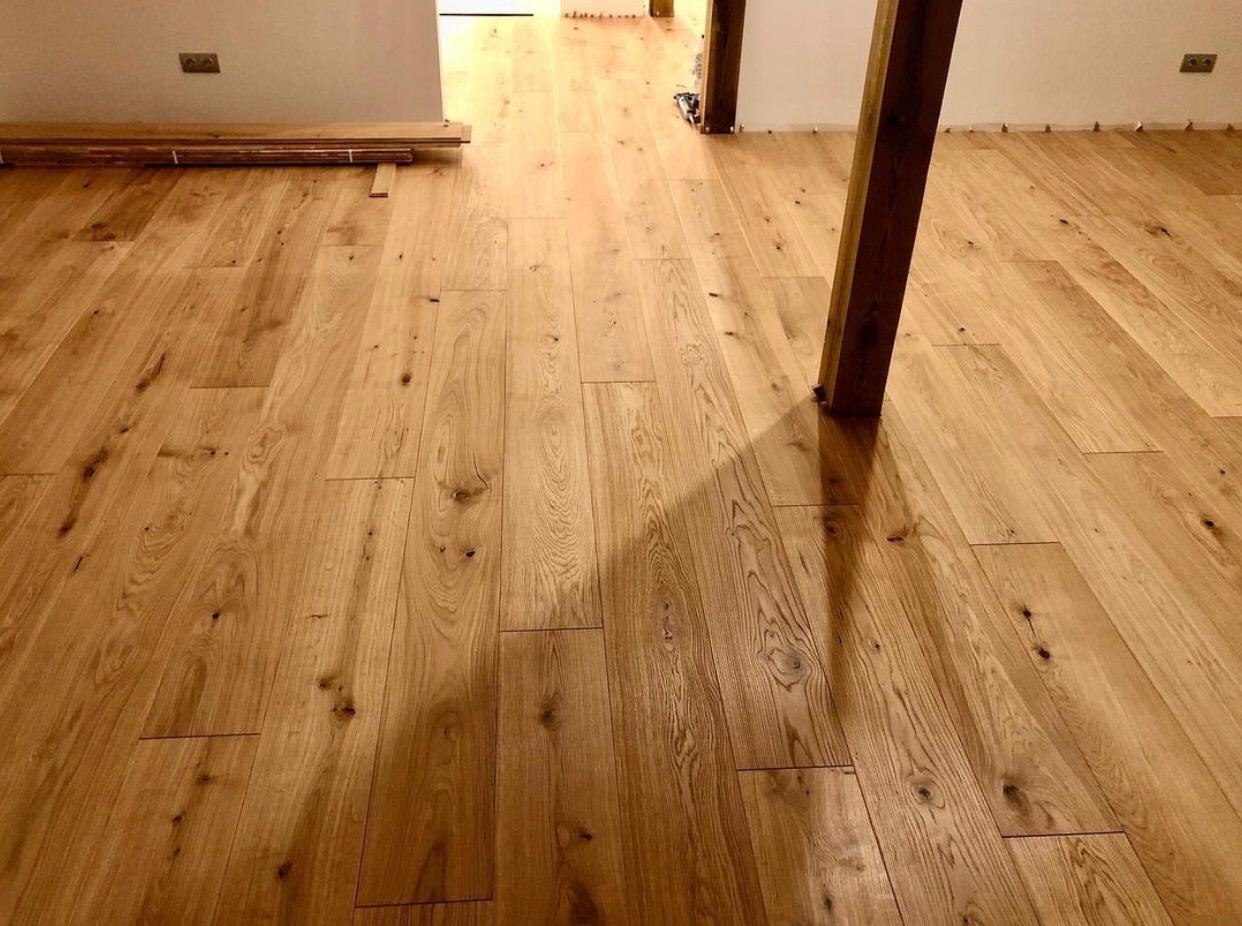 Jasny, drewniany parkiet ułożony w przestronnym wnętrzu, z widocznymi słupami konstrukcyjnymi i stos paneli w tle, oświetlony ciepłym światłem.