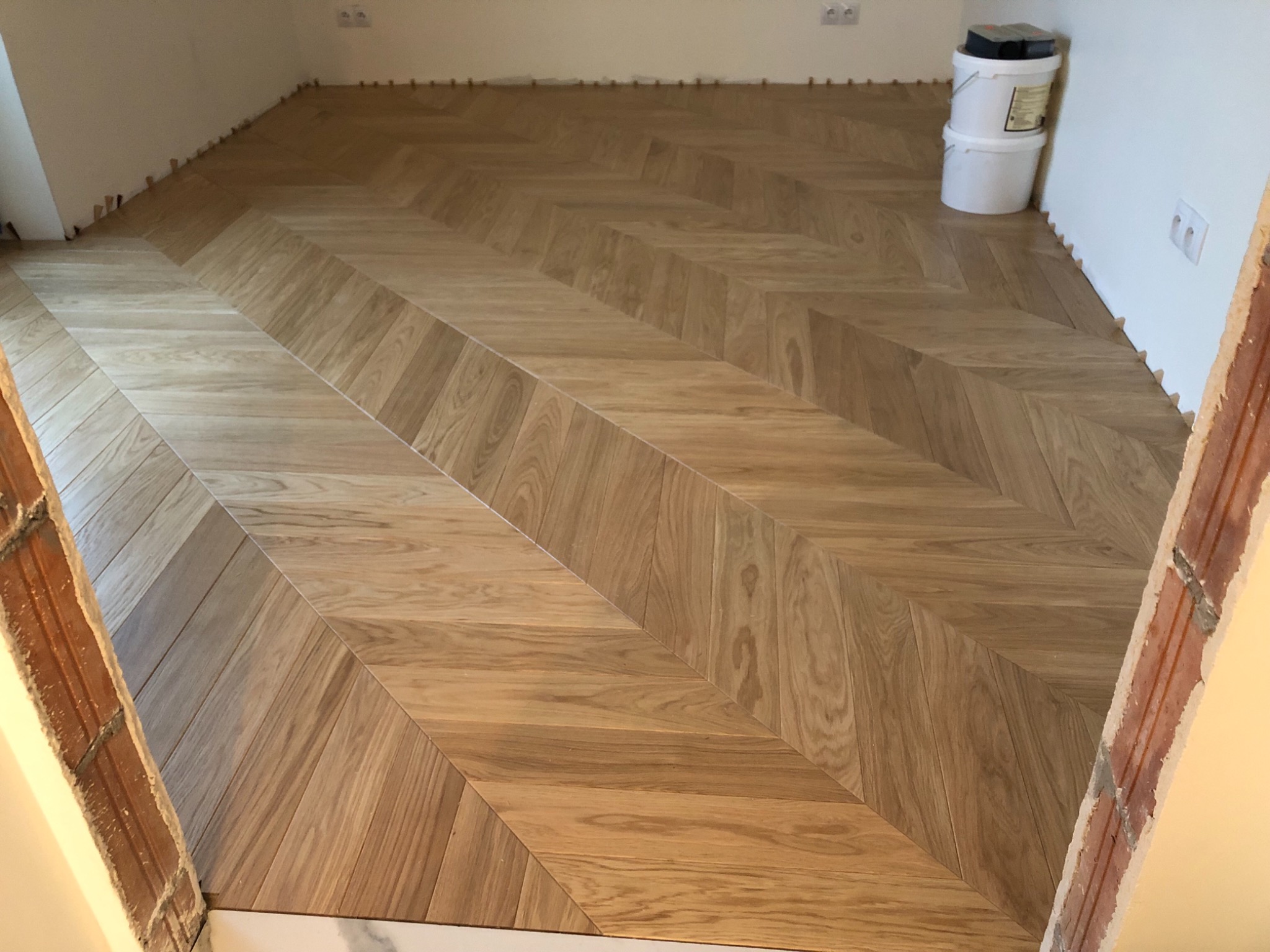 Jasny parkiet ułożony we wzór jodełki w trakcie montażu, widoczne kliny dystansowe przy ścianach i dwa białe wiadra z zaprawą w rogu pomieszczenia.