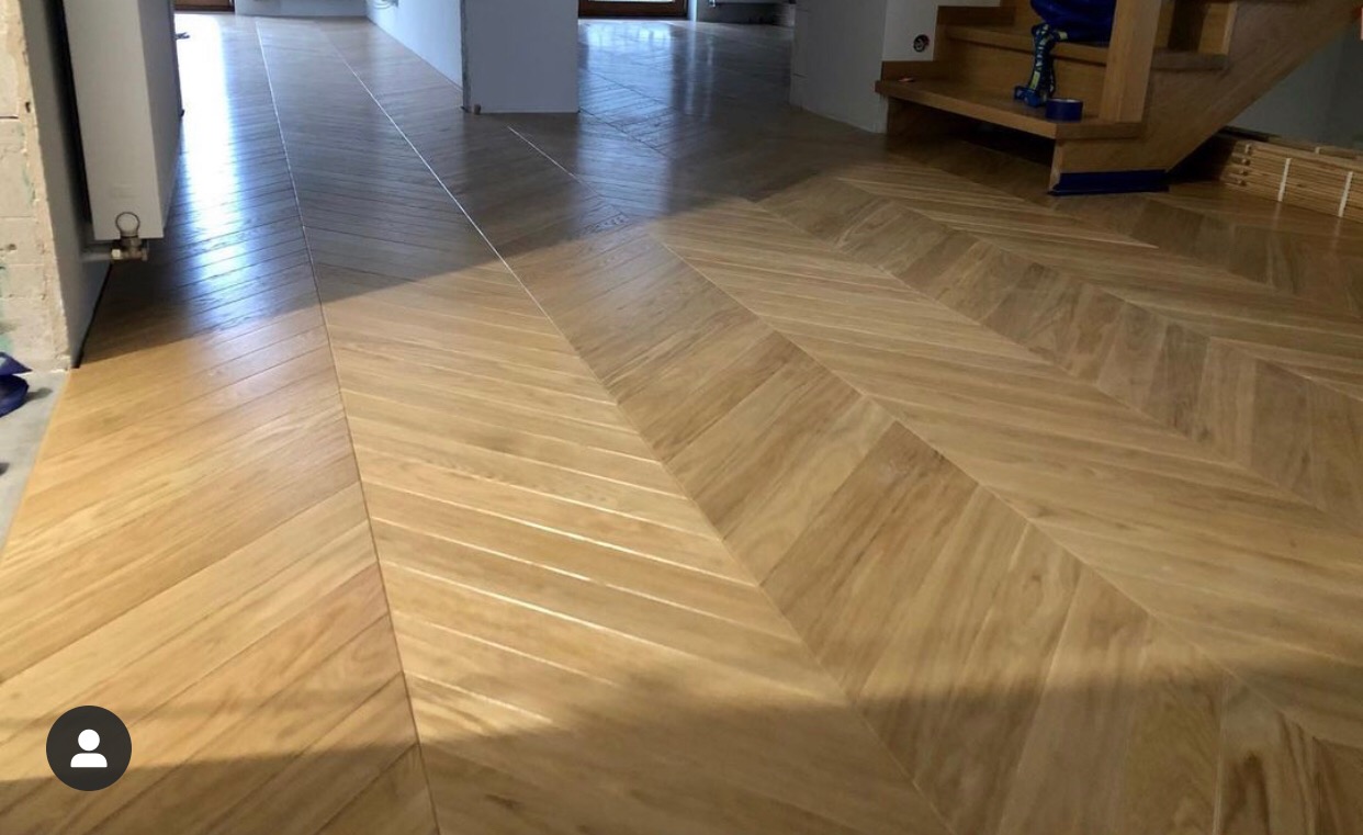 Jasny parkiet ułożony we wzór jodełki francuskiej, widoczne niedokończone prace wykończeniowe przy ścianie i schody w tle.