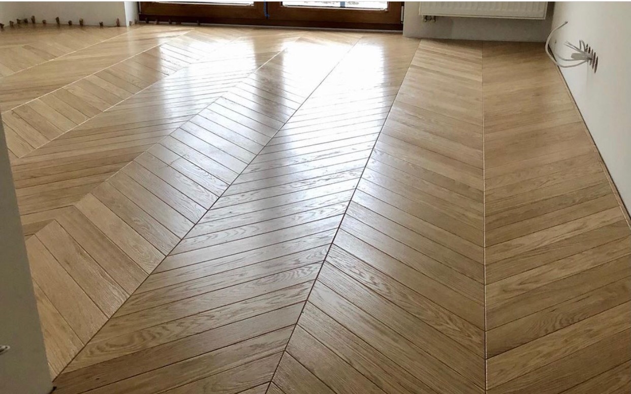 Jasny parkiet ułożony we wzór jodełki francuskiej, z widocznymi detalami usłojenia drewna i odbiciem światła od okna.