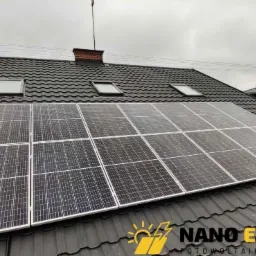 Panel słoneczny na ciemnym dachu z dachówką, widoczne okna dachowe i komin z cegły, pochmurne niebo w tle.