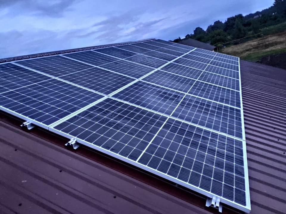 Panel słoneczny na brązowym dachu z blachy trapezowej, widoczne mocowania, pochmurne niebo w tle.