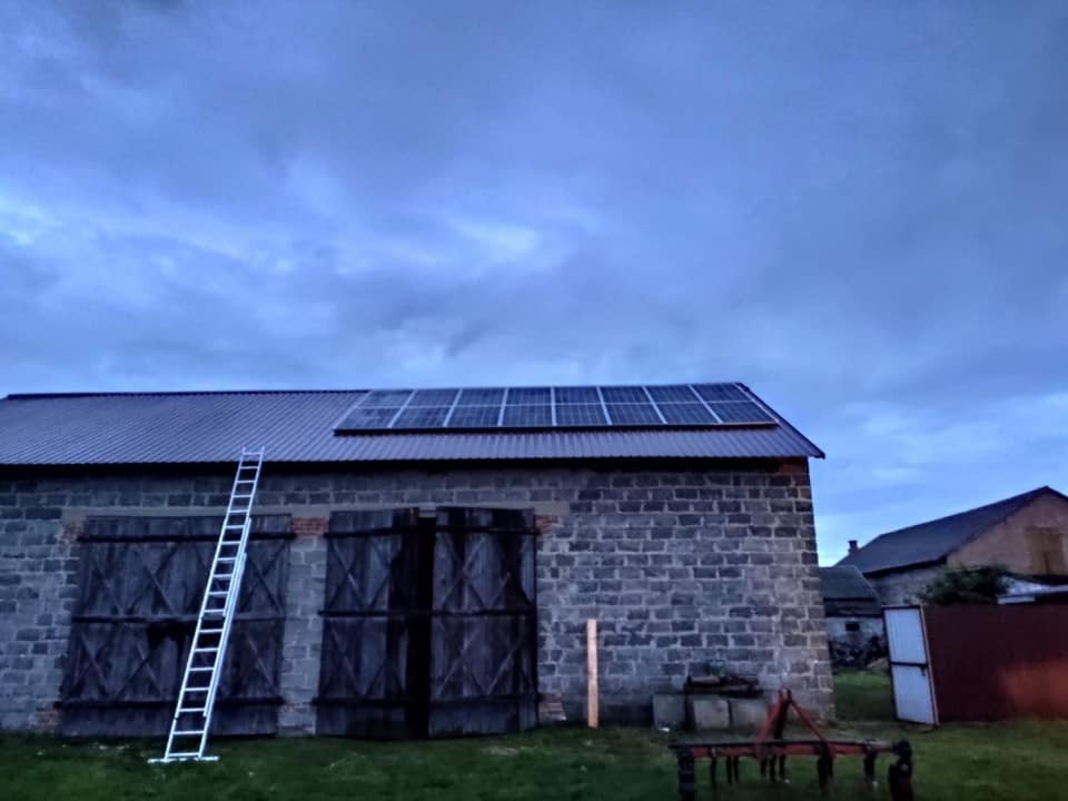 Instalacja fotowoltaiczna na dachu stodoły z cegły, drabina oparta o ścianę, pochmurne niebo.